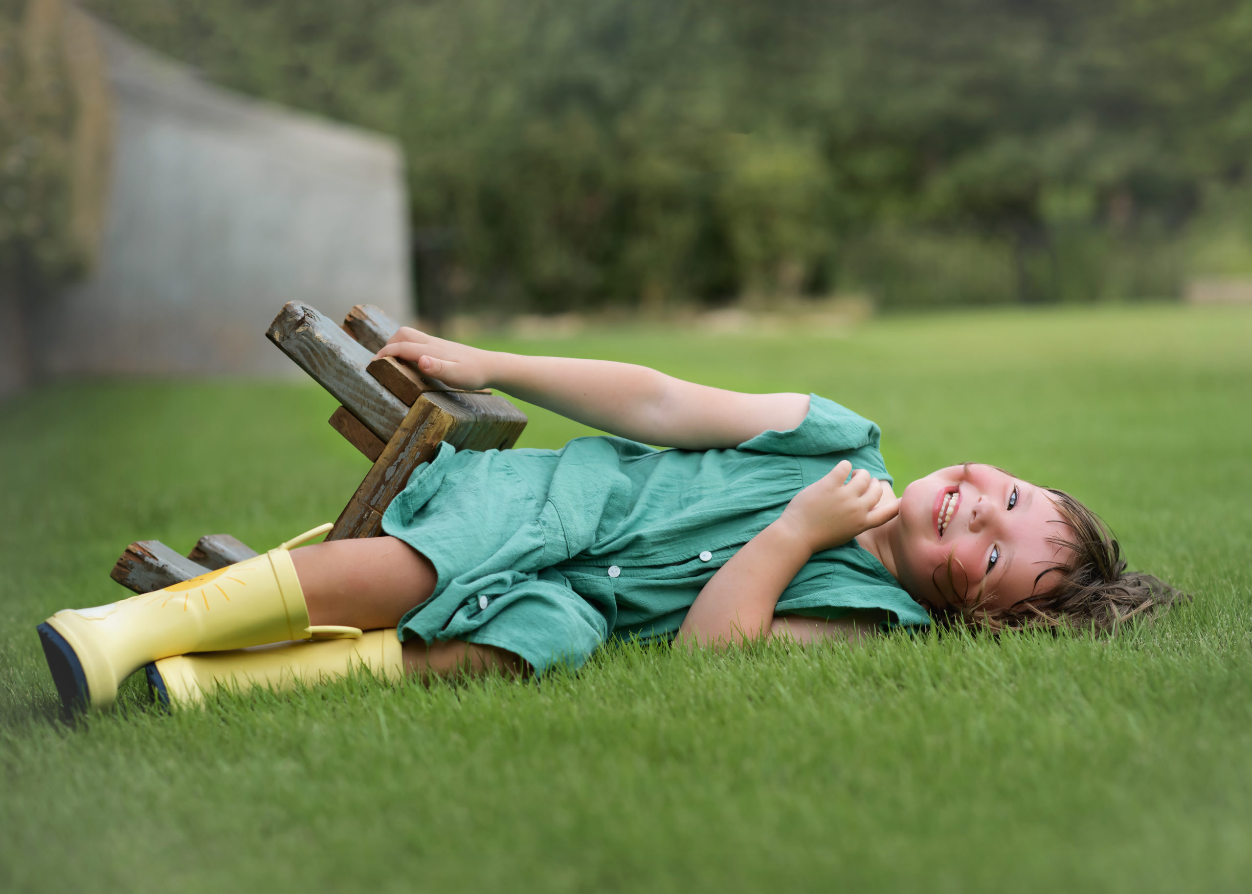 little girl in green dress and yellow rainboots laughing and laying on ground after tipping over the stool she was sitting on