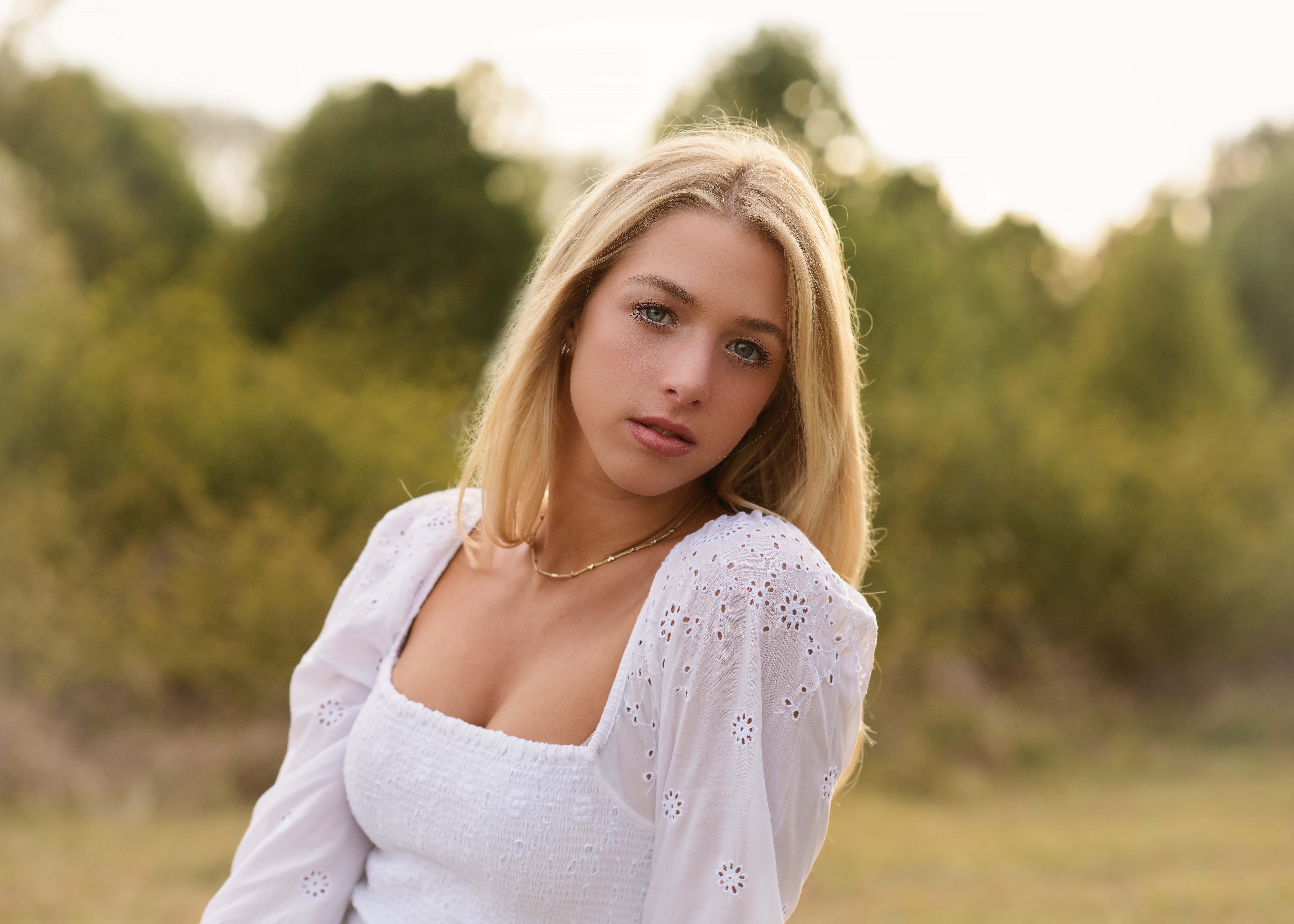 Young blonde girl outdoors in front of green trees in white peasant type top slightly looking over her shoulder with a serious face