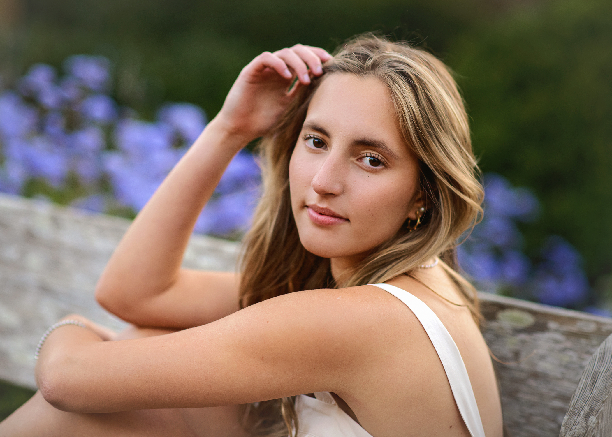 Senior girl sitting on wooden bench in white dress with soft smile and purple flowers in the background