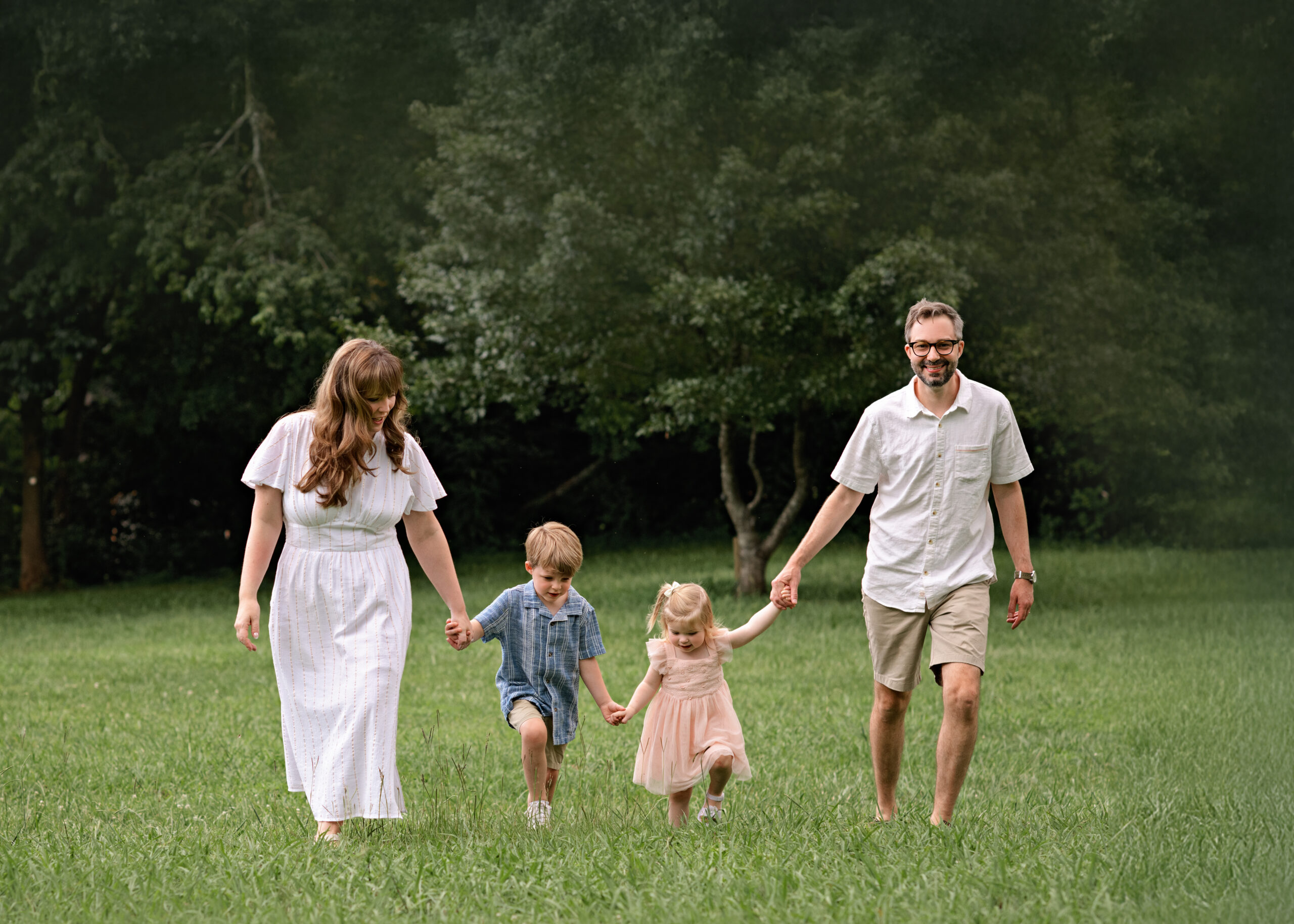 Family of four during an outdoor photo session with a lifestyle family photographer in Atlanta, Georgia. Parents walk hand-in-hand with their young children in a grassy field, surrounded by trees. Candid, natural light family photography